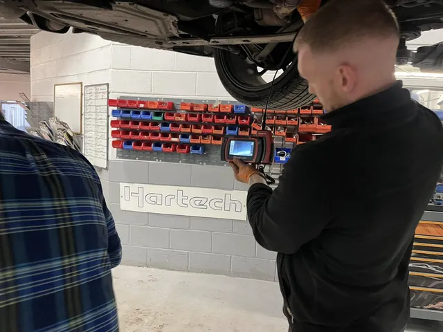 A technician under a Porsche on a lift, studying a borescope screen, with the Hartech name painted on the workshop wall behind.