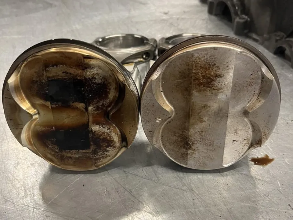 Two pistons side by side on the bench, one crown heavily coated in carbon.