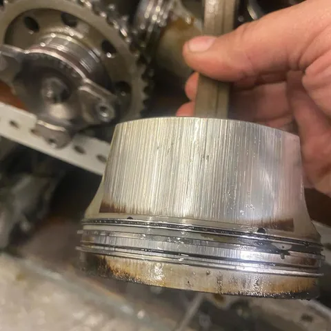 A technician holding a piston with a deeply scored skirt, with the timing chain and sprockets behind.