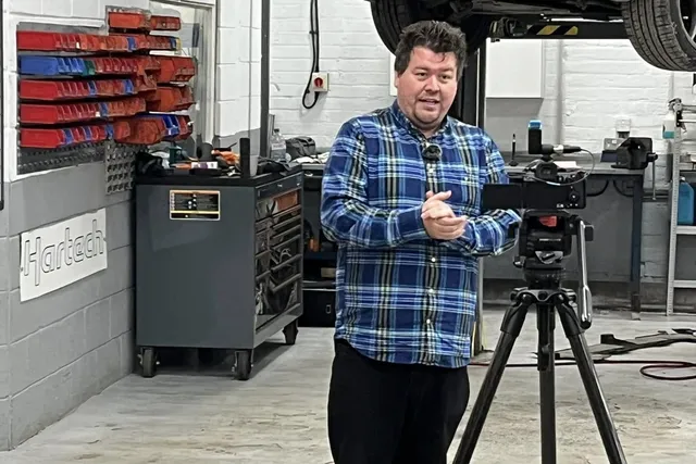 A man with a video camera on a tripod in the workshop, a Boxster raised on the lift behind him.