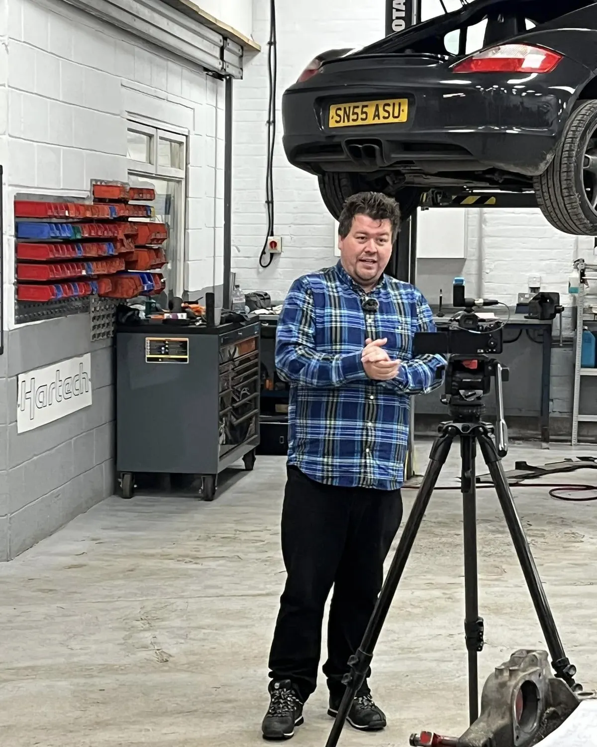 A man with a video camera on a tripod in the workshop, a Boxster raised on the lift behind him.