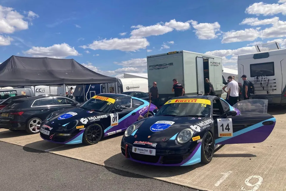 Race-liveried 911s parked in the paddock in front of awnings and transporters.