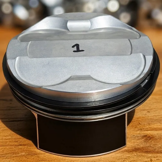 A single new forged piston marked with the number 1, standing on a wooden bench.