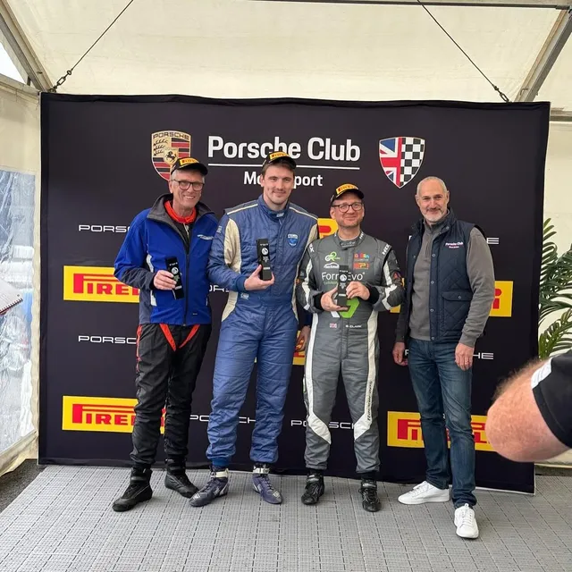 Four people on a Porsche Club Motorsport podium backdrop, two in race suits holding trophies.