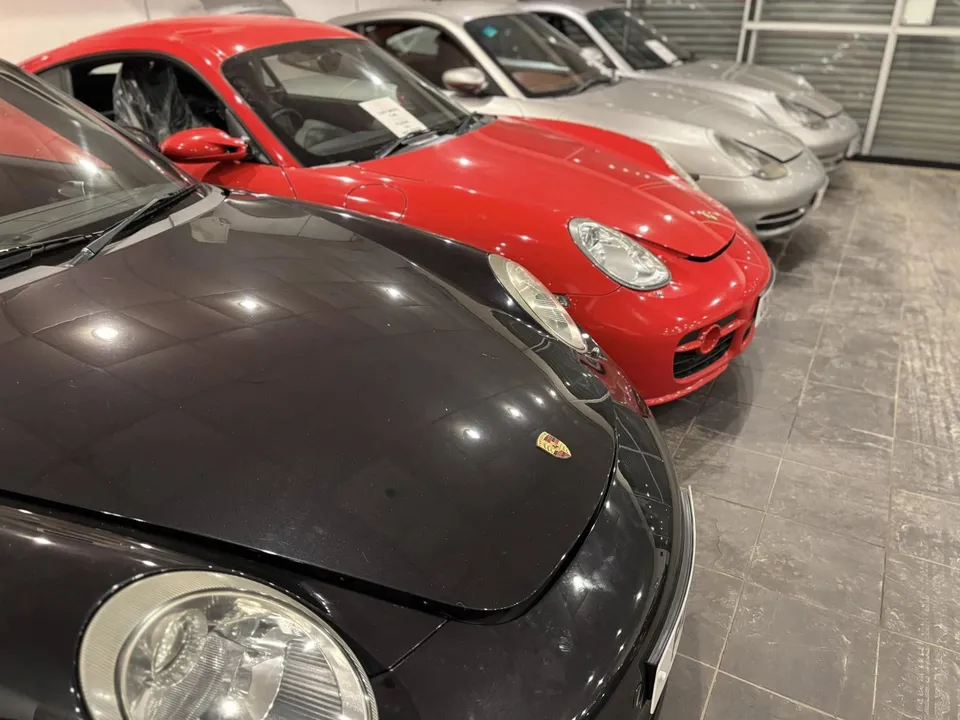 Several Porsches parked close together indoors under the lights: a dark 997 nearest, a red Cayman behind it.