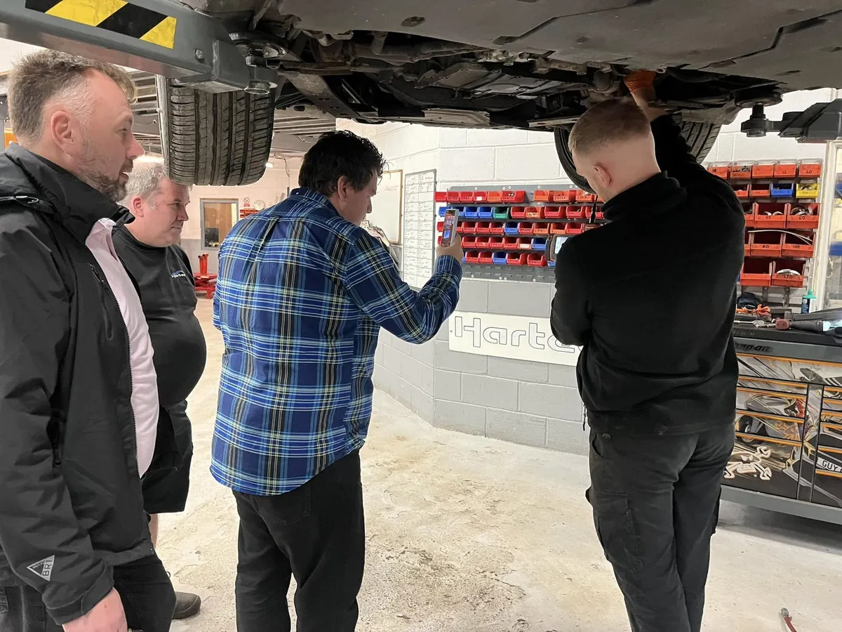 Two technicians and two visitors standing under a raised Porsche, looking up at the underside together.