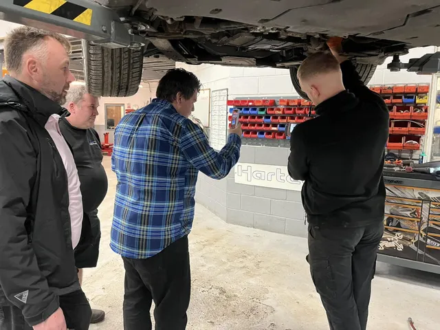 Two technicians and two visitors standing under a raised Porsche, looking up at the underside together.